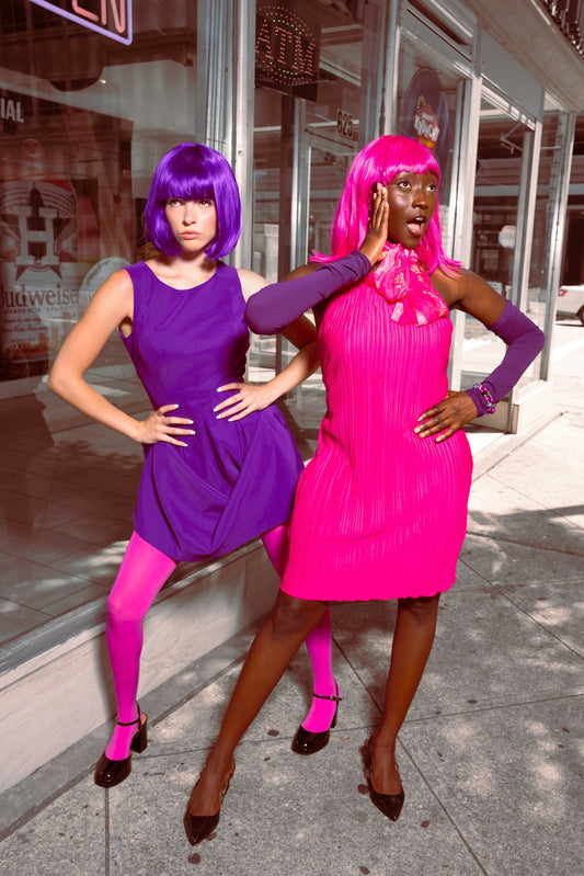 Two women in vibrant purple and pink outfits and wigs.