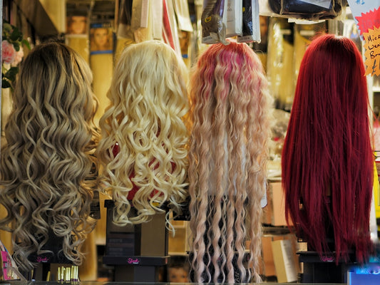 Four wigs in blonde, platinum, pink, and red displayed on stands in a shop